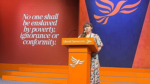 Katharine Macy at the Conference podium in a hall with bold coloured panels and a Lib Dem logo behind her